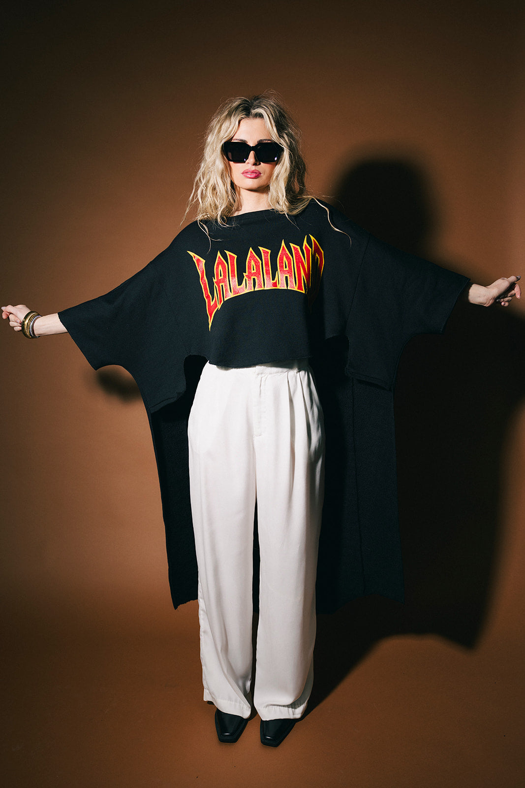 Person wearing a black oversized sweatshirt with 'LALALAND' text, white pants, and sunglasses on a brown background