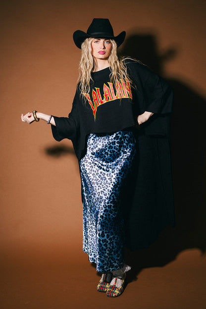 Person wearing a black oversized sweater with orange text, blue leopard print pants, and a black cowboy hat against a brown background.