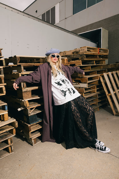 Person standing among wooden pallets wearing a plaid coat, graphic t-shirt, and sunglasses.