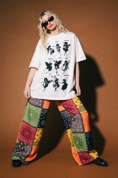 Person wearing a white t-shirt with black graphics and colorful patchwork pants on a brown background