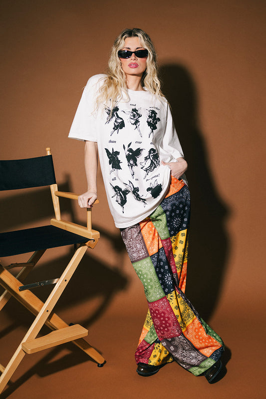 Person wearing a white graphic t-shirt and colorful patchwork pants standing next to a director's chair on a brown background.