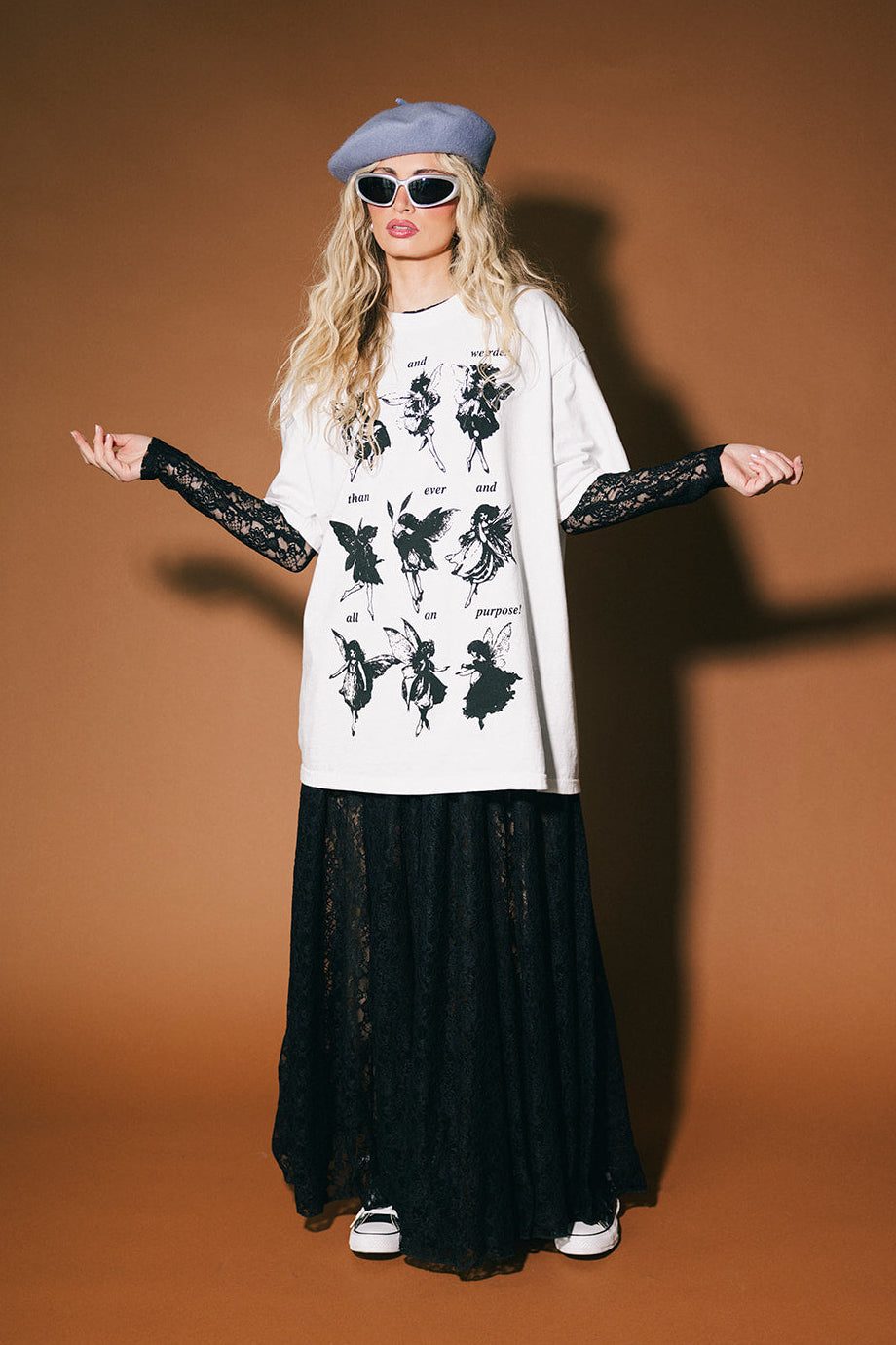 Person wearing a white graphic t-shirt with black designs, black long-sleeve shirt underneath, black skirt, sunglasses, and beret on a brown background.