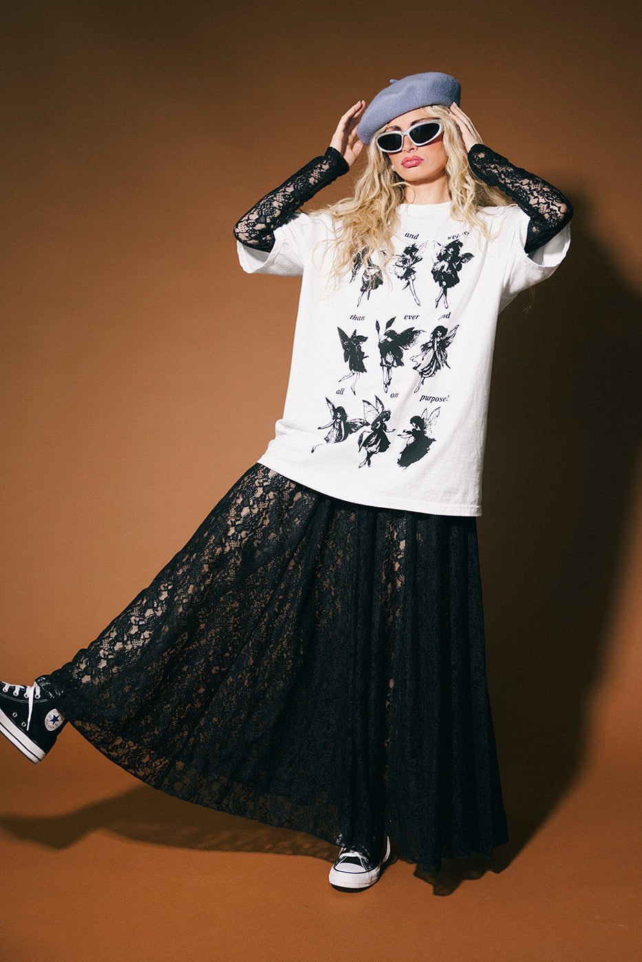 Person wearing a white graphic t-shirt, black lace top, black lace skirt, and sneakers on a brown background