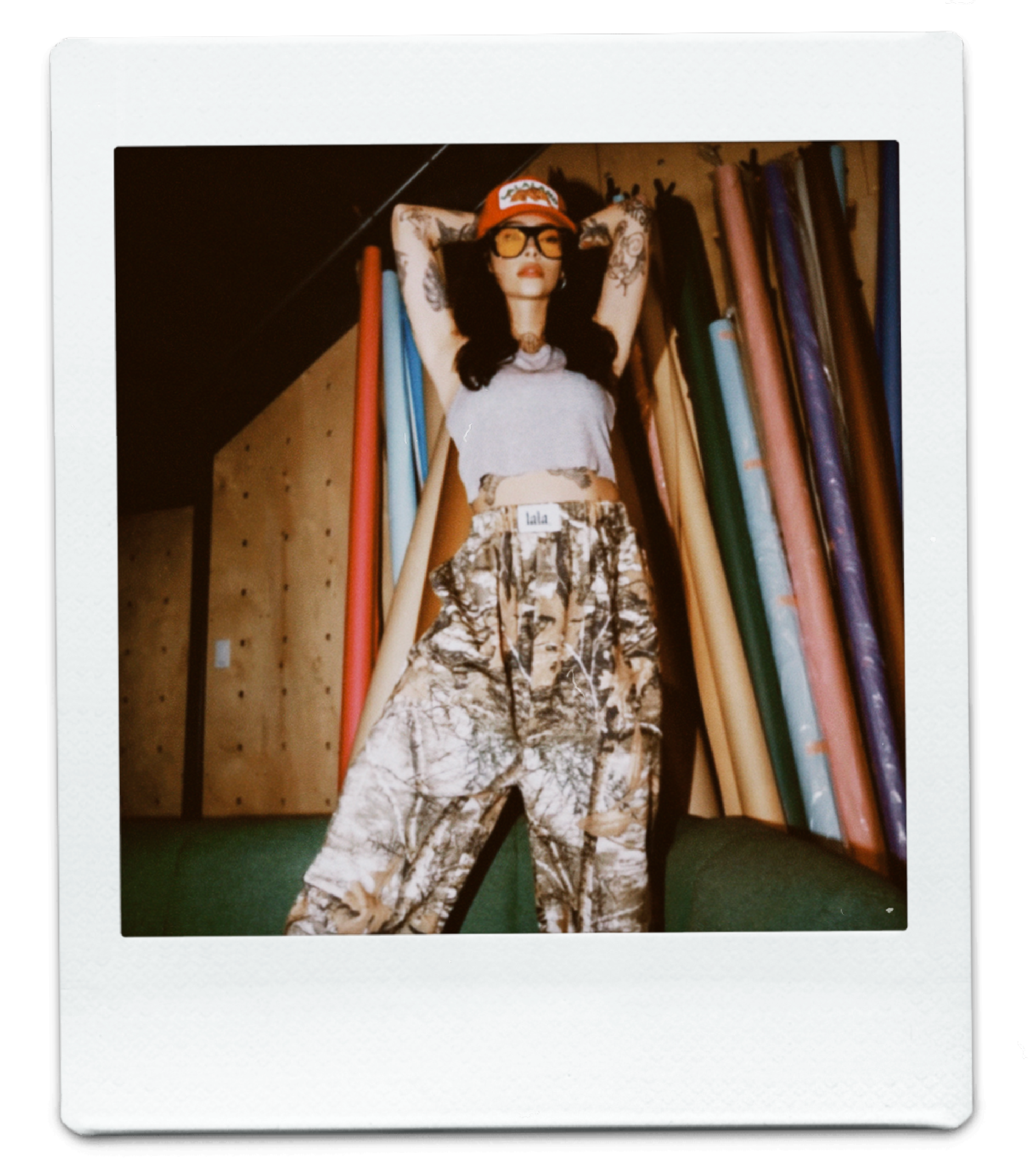 Polaroid photo of a lala model wearing a red trucker hat, grey tank top, and camo pants