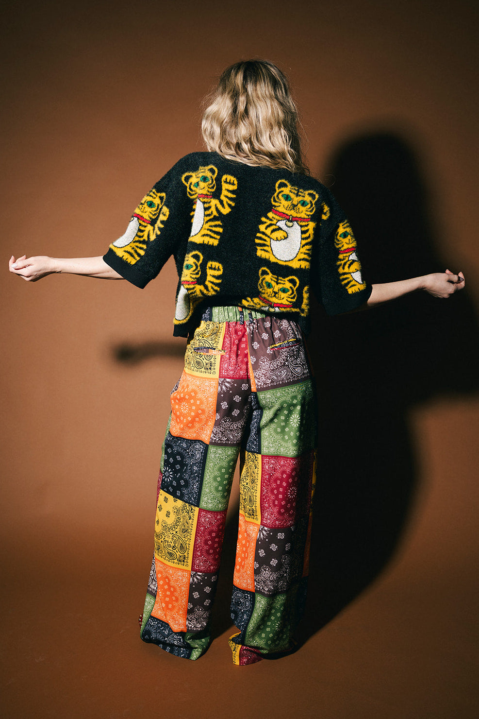 Person wearing a colorful outfit with a patterned jacket and wide-leg pants on a brown background