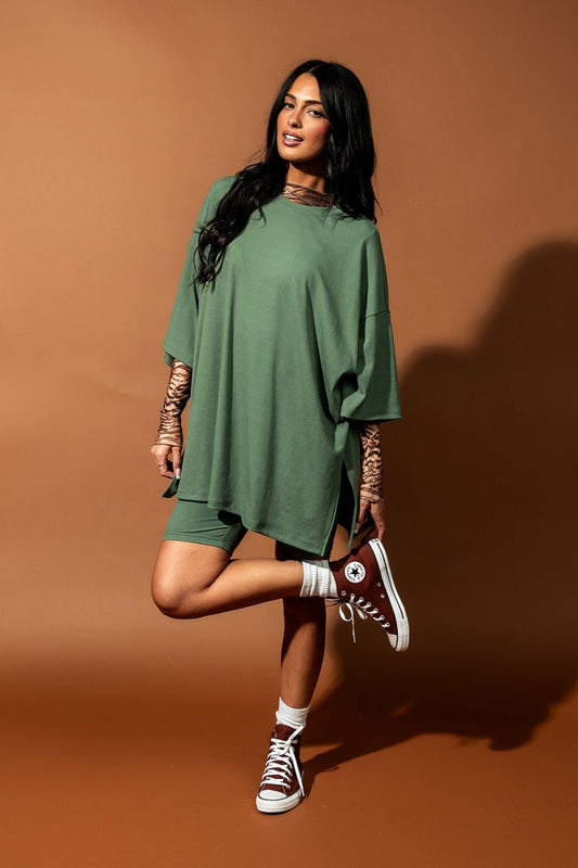 Girl/model standing against a brown backdrop wearing a sage green biker set with a tiger striped mesh top underneath