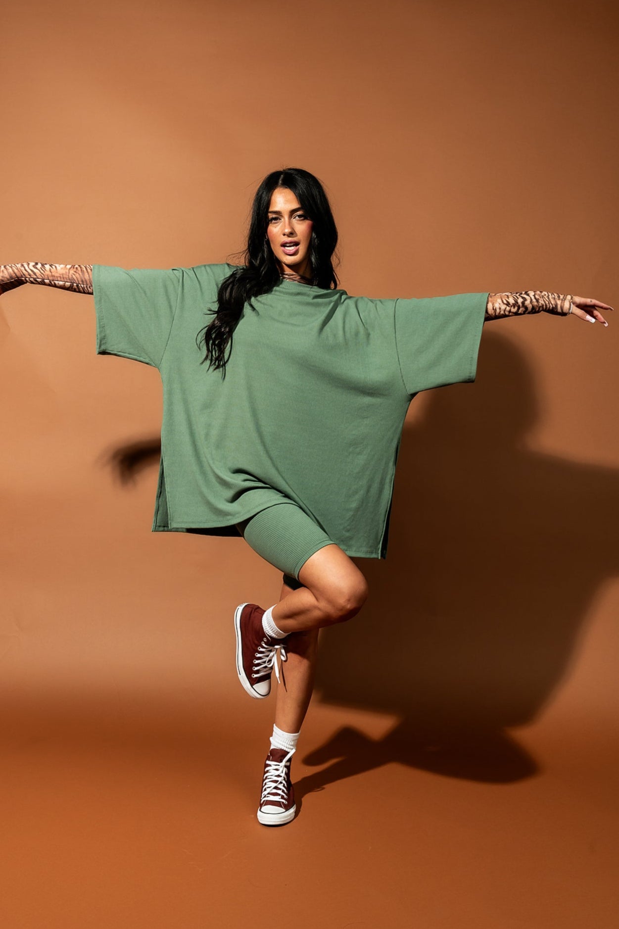 Girl/model standing against a brown backdrop wearing a sage green biker set with a tiger striped mesh top underneath