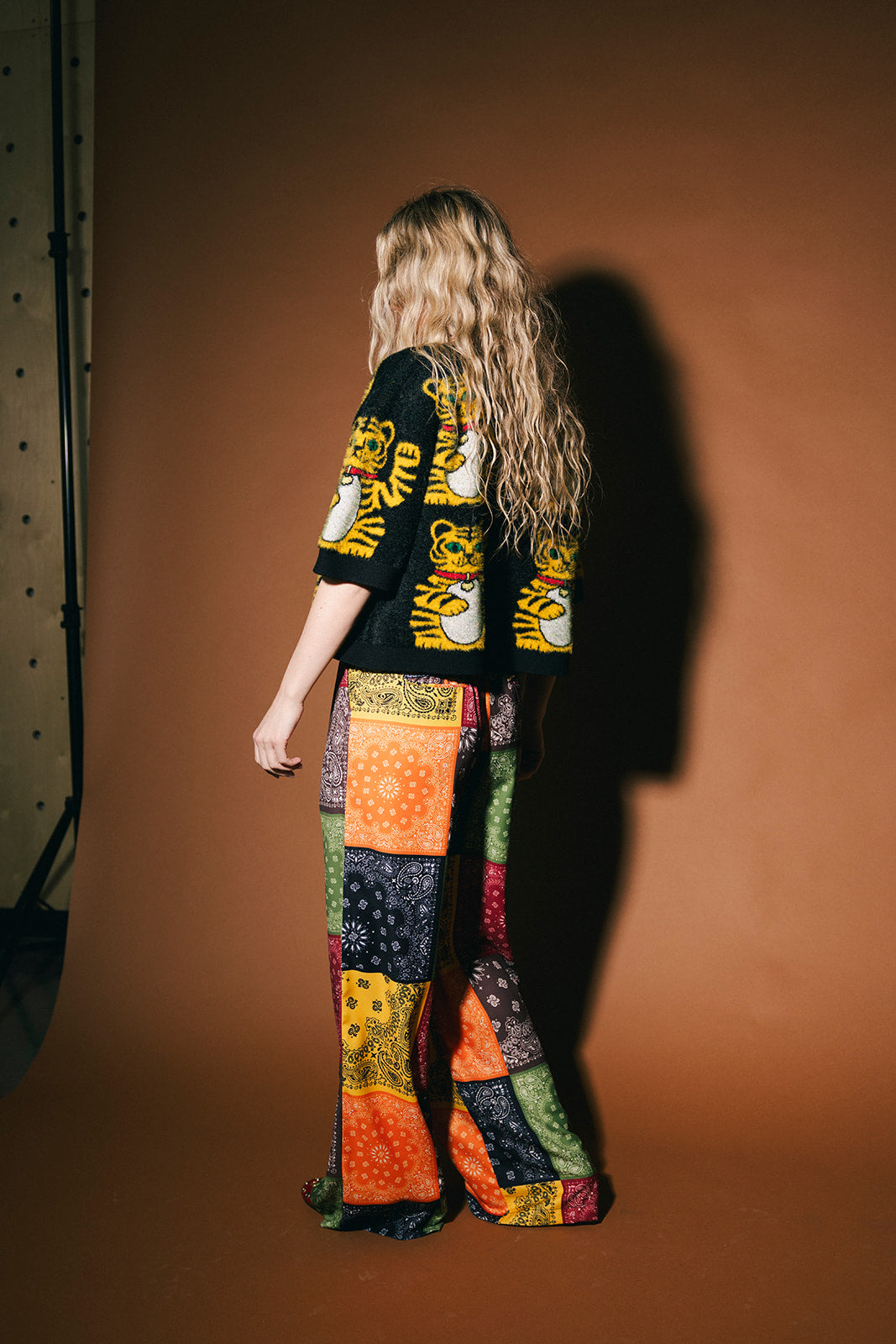 Person wearing a black graphic t-shirt and colorful patchwork pants against a brown background