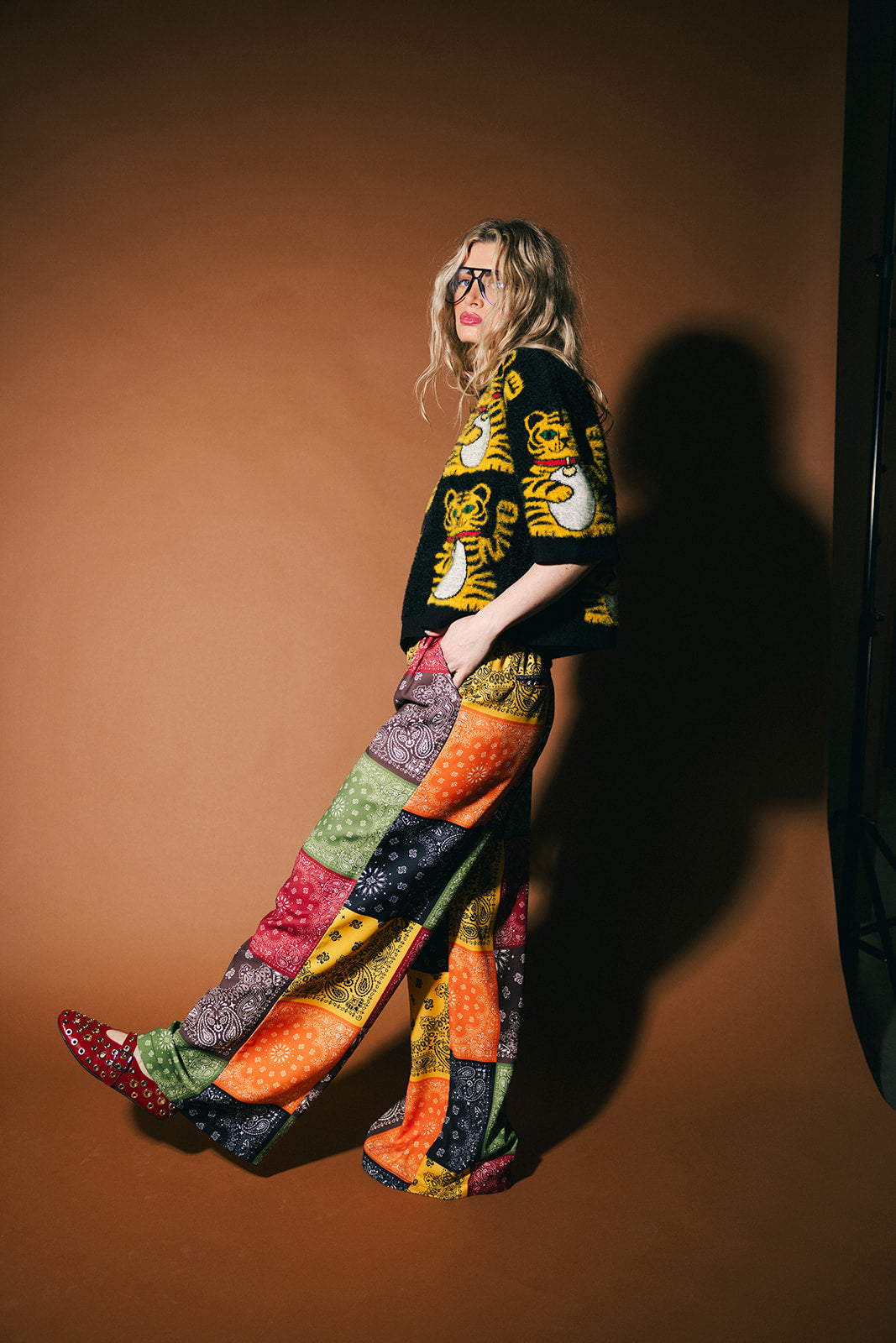 Person wearing a black graphic t-shirt and colorful patchwork pants against a brown background