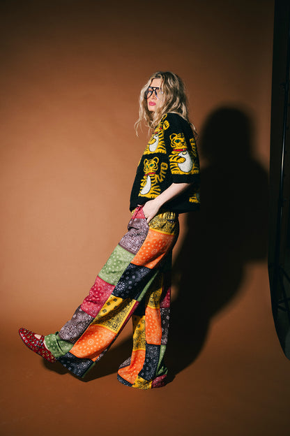 Person wearing a black graphic t-shirt and colorful patchwork pants against a brown background