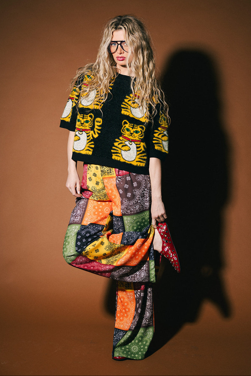 Person wearing a colorful outfit with a patterned shirt and patchwork skirt against a brown background
