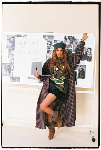 Person in a plaid duster and green beret posing in front of a wall with black and white photos.