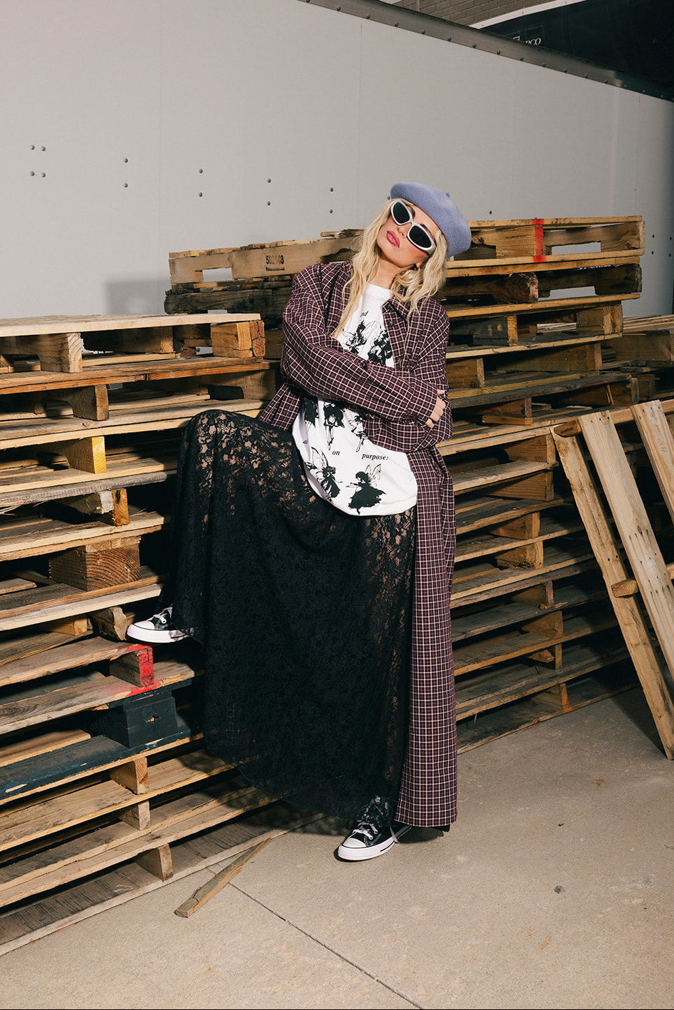Person standing in a warehouse with wooden pallets, wearing a plaid duster and black skirt.