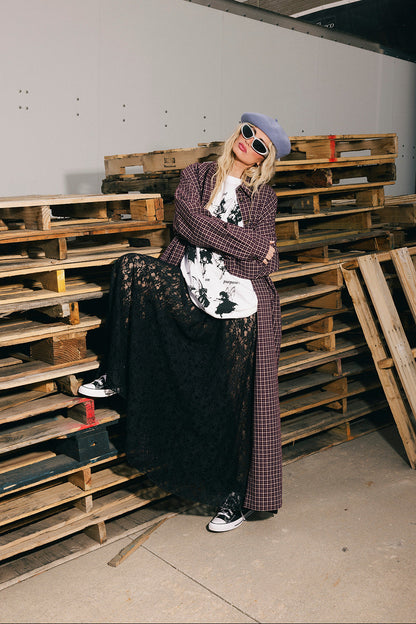 Person standing in a warehouse with wooden pallets, wearing a plaid duster and black skirt.
