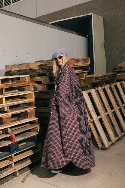 Person wearing a long plaid duster with text, standing in an industrial setting with wooden pallets.