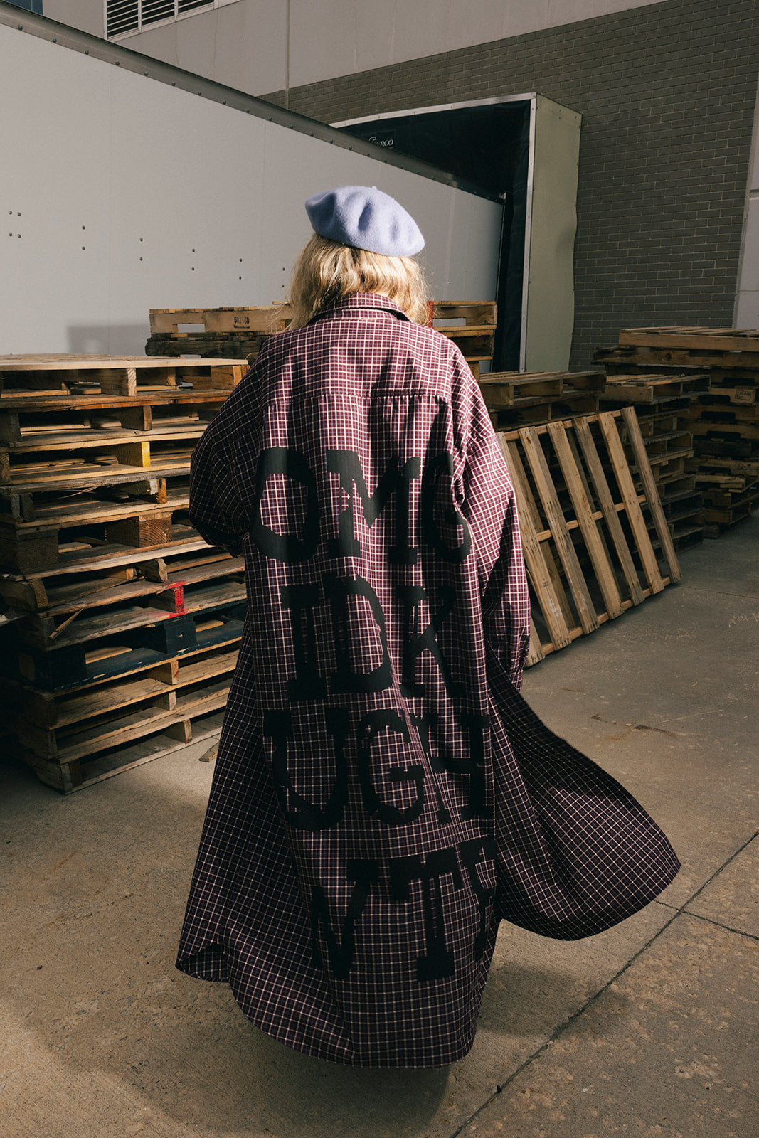 Person wearing a long, checkered duster with text, standing in an industrial setting with wooden pallets.