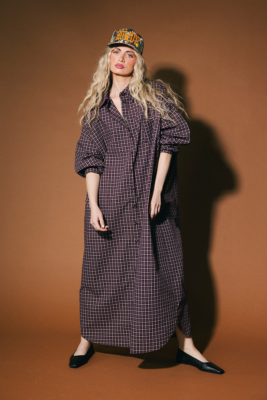 Woman wearing a long plaid duster and cap against a brown background