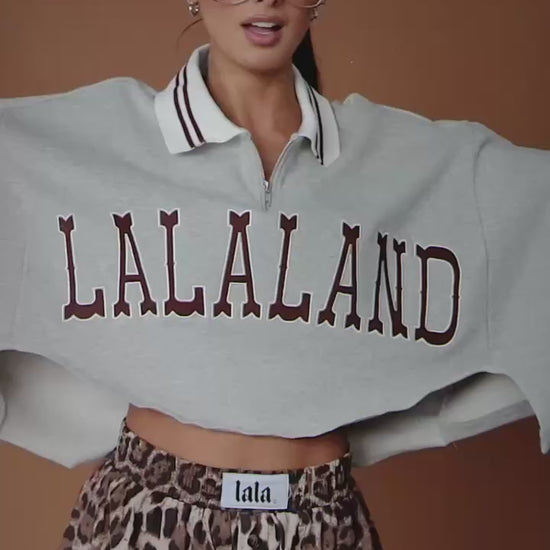 Video of woman wearing a grey cropped polo with "LALALAND" text against a brown backdrop.