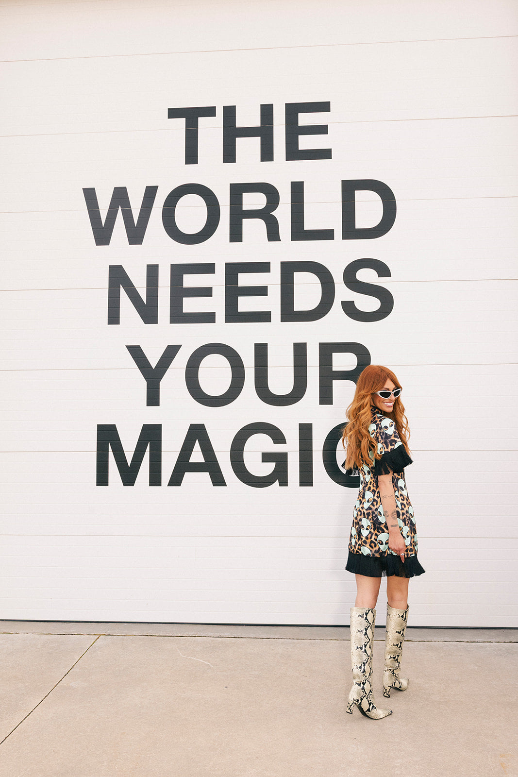 Woman in a floral dress standing in front of a wall with 'The World Needs Your Magic' text.