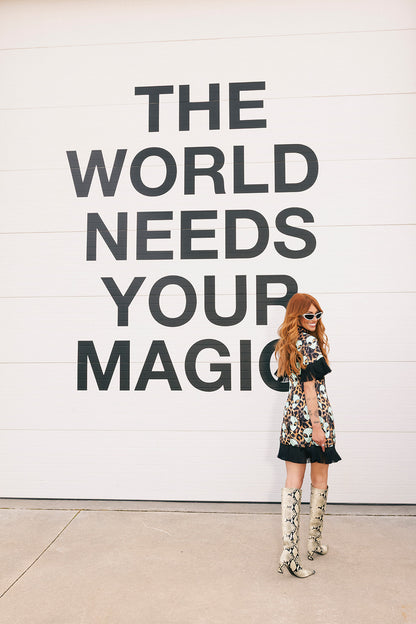 Woman in a floral dress standing in front of a wall with 'The World Needs Your Magic' text.