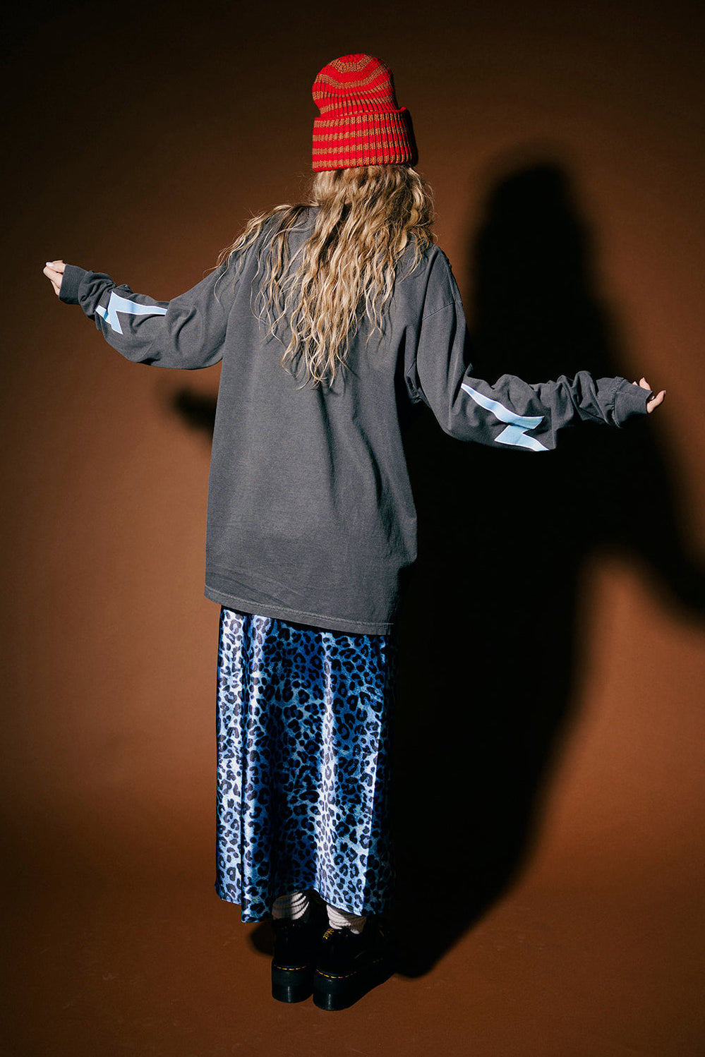 Person wearing a red beanie and gray hoodie with arms outstretched on a brown background